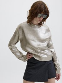 Μεταλλική χρυσή μπλούζα με υφή, στρογγυλή λαιμόκοψη, μακρυά μανίκια, συνδυασμένη με μια σκοτεινή, κοντή φούστα που διαθέτει πλευρική τσέπη.