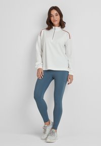 Woman stands against white background wearing white zip-up sweatshirt, blue leggings, and light gray sneakers.