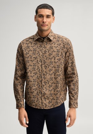 Uomo che indossa una camicia marrone con bottoni e motivo floreale nero, in piedi con le braccia rilassate, su uno sfondo grigio chiaro uniforme.