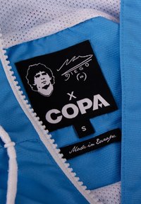Blauwe sporttrui met een mesh interieur, witte rits en merklabel met grafische elementen. Het label bevat "COPA," "Gemaakt in Europa," en maat "S."