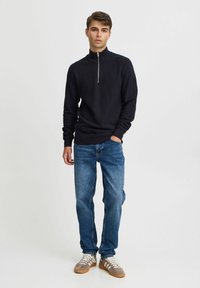 Pulover cu fermoar la gât de culoare bleumarin, dintr-un material texturat, asortat cu blugi de culoare albastru deschis și adidași gri cu șireturi albe și accente bej.