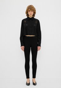 Schwarzes, gekürztes Shirt mit gesticktem Detail auf der Brust, kombiniert mit schwarzen Leggings und schwarzen Ballerinas, was ein monochromatisches Outfit ergibt.
