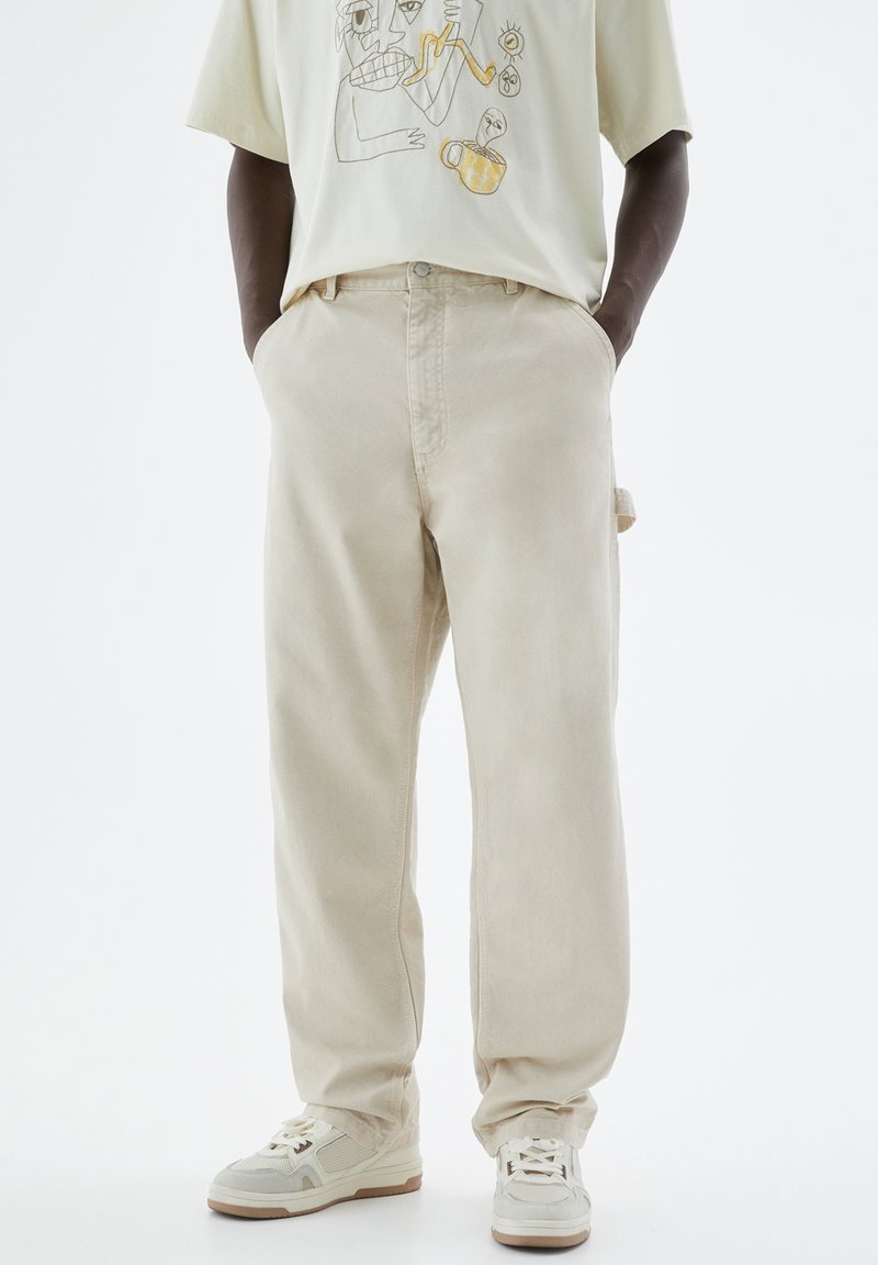 Homme portant un pantalon beige ample, des baskets blanches et beiges, et un t-shirt beige clair avec des imprimés abstraits de visages et d'objets. Mains dans les poches.