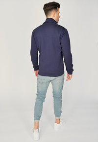 Granatowy sweter z wysokim kołnierzem, długimi rękawami i prążkowanymi mankietami, w połączeniu z jasnoniebieskimi, obcisłymi dżinsami i białymi trampkami.