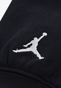 Černá tkanina s texturovaným povrchem, na které je vyšitý bílý logo Jumpman, znázorňující skákající postavu držící basketbal.