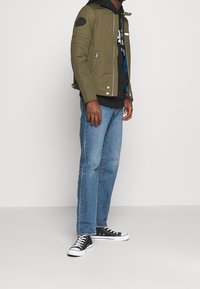 Man som bär en olivgrön zip-up jacka, svart hoodie, blå jeans och svarta högstövlar, stående mot en vit bakgrund.