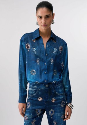 Donna che indossa una camicia blu e jeans abbinati con motivi floreali e cammeo, orecchini d'argento e un braccialetto d'argento, in piedi davanti a uno sfondo semplice.