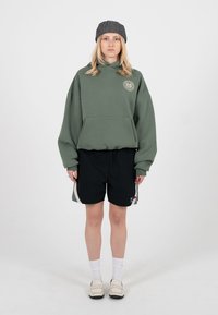 Blond inimene seisab, seljas hall müts, liiga suur roheline kapuutsiga dressipluus logoga, mustad spordipüksid, valged sokid ja beežid lamedad kingad valgel taustal.