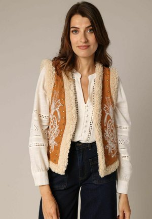 Bruine vest met decoratieve witte borduursels en schapenvachtafwerking, gedragen over een witte blouse met gaatjesdetails. Gemaakt met donkere jeans.