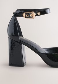Black patent leather heel featuring a block shape, ankle strap with gold buckle, and a smooth texture with shiny finish.