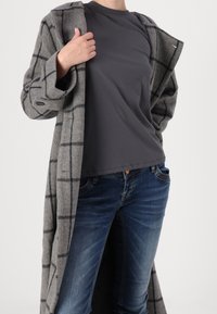 Grå ullkappa med svart rutmönster, kombinerad med en mörk t-shirt och blå jeans. Kappan har en avslappnad passform och stora fickor.