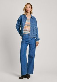 Giubbotto di denim con motivo trapuntato, abbinato a un maglione in maglia multicolore e jeans a gamba larga blu. Stivali neri alla caviglia completano l'outfit.