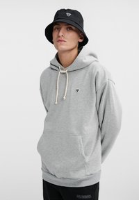 Grå hoodie med en framficka, dragsko i huvan och liten svart logotyp på bröstet. Matchad med en svart bucket hat. Slät tygtextur.