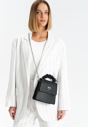 Borsa nera con texture, manico in stile scrunchie e tracolla a catena, dotata di una patta frontale e logo argentato, indossata con un blazer bianco a righe.