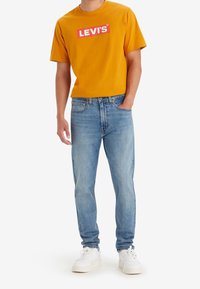 Gul kortärmad t-shirt med röd "LEVI'S" logotyp, tillsammans med ljusblå jeans och vita sneakers. Avslappnad passform, standard denimdesign.