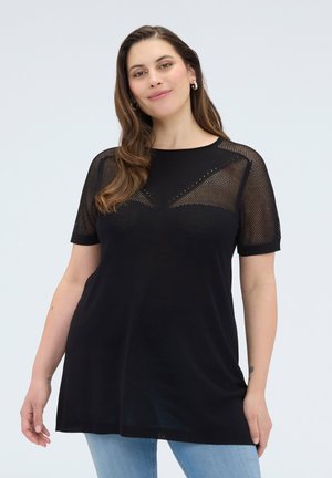 Kvinde med langt brunt hår iført guld øreringe, en sort kortærmet mesh-top og lyseblå jeans, stående mod en ensfarvet baggrund.