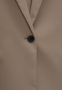 Blazer beige en tissu texturé, doté d'un unique bouton noir et d'un col à revers. Fini lisse avec des détails de couture subtils.