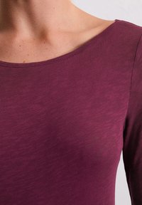 Langärmliges Oberteil in tiefem Burgunderrot, aus weichem, strukturiertem Stoff. Verfügt über einen weiten Rundhals und einen subtilen Glanz.