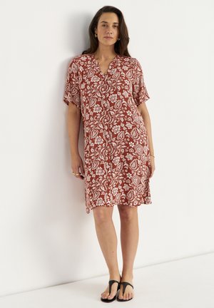 Kvinde stående mod en hvid væg iført en knælange, kortærmet rustfarvet kjole med hvide blomsterprint og sorte sandaler.