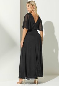 Vestido de noche negro con un diseño de espalda en V, mangas acampanadas y una falda fluida. Presenta una sutil textura brillante. Llevado con tacones dorados.