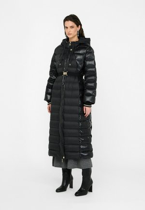 Femme portant un long manteau en duvet noir ceinturé avec capuche, une jupe grise, des bottes à talons noires et des boucles d'oreilles pendantes, se tenant contre un fond blanc.
