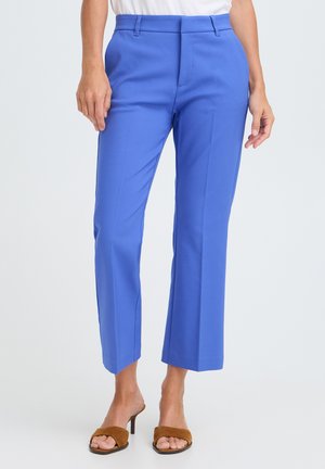 Vrouw draagt felblauwe cropped broek met bruine open teen sandalen met hak en een wit topje, staand tegen een effen achtergrond.