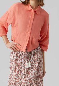 Camisa coral de botones con una superficie texturada y mangas sueltas de media longitud, combinada con una falda de estampado floral que presenta adornos de borlas.