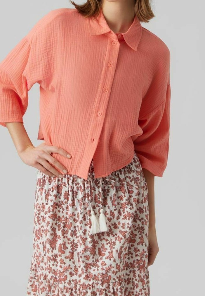 Camisa coral de botones con una superficie texturada y mangas sueltas de media longitud, combinada con una falda de estampado floral que presenta adornos de borlas.