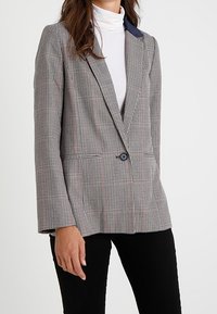 Blazer s houndstooth vzorem v béžové a černé barvě, s námořnickým límcem, jednobotonovým zapínáním a strukturovaným střihem s dlouhými rukávy.