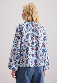 Donna bionda che indossa una blusa bianca con un motivo floreale blu e rosso e pantaloni larghi a vita alta blu, rivolta a sinistra su uno sfondo chiaro.