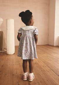 Blaues Blumenkleid mit kurzen Ärmeln und einem Spitzkragen, kombiniert mit pinken Turnschuhen und weißen, mit Rüschen verzierten Socken. Hintergrund aus Holzboden.