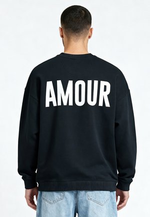 Man met kort donker haar draagt een zwart sweatshirt met grote witte "AMOUR" tekst op de rug en lichtblauwe spijkerbroek, staand tegen een witte achtergrond.