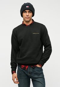 Μαύρο φούτερ με ριγέ μανίκια και λαιμόκοψη, που φέρει ένα μικρό χρυσό λογότυπο. Φορεμένο πάνω από ένα καρό πουκάμισο σε κόκκινο και μαύρο χρώμα και μπλε τζιν.