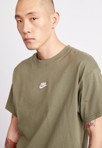 Olīvu zaļa koka t-kreklis ar īsām piedurknēm, ar baltu Nike logotipu uz krūtīm. Gluda tekstūra un klasiskā apļa izgriezuma forma.