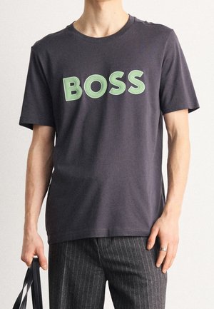 T-shirt di cotone grigio scuro con un grande logo verde "BOSS". Maniche corte, scollo rotondo, abbinata a pantaloni a righe, tenuta da una mano che porta una borsa.