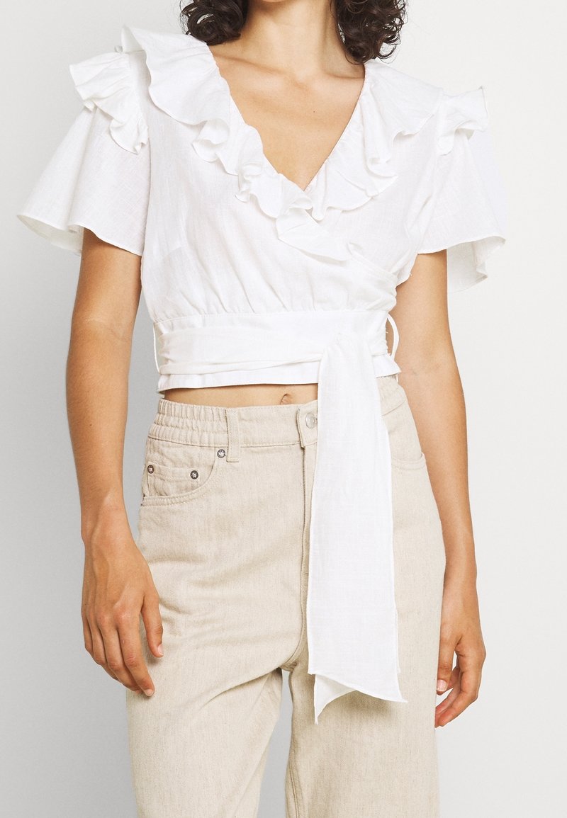Femme portant un crop top blanc cache-cœur avec des manches à volants et un pantalon beige taille haute avec une ceinture élastique.
