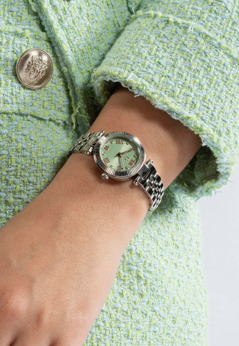 Orologio da polso in tonalità argento con lunetta zigrinata, quadrante verde menta, numeri romani e bracciale metallico indossato su una mano su un tessuto verde texturizzato.