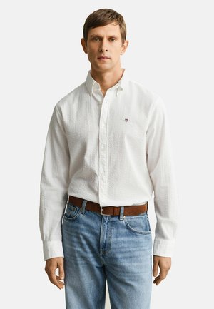 Mann trägt ein strukturiertes weißes Hemd mit Knöpfen, das in eine hellblaue Jeans mit braunem Gürtel gesteckt ist, und steht vor einem schlichten Hintergrund.