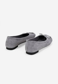 Un paio di ballerine in suede grigio con piccoli fiocchi sulla parte superiore e suole nere piatte, viste da un angolo posteriore su sfondo bianco.