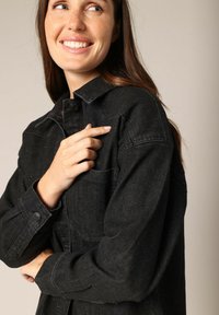 Zwarte denimjas met een knoopsluiting aan de voorkant, een borstzak en lange mouwen. Textuurmateriaal met subtiele spikkels erdoorheen.