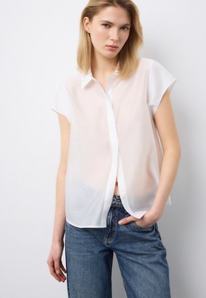 Junge Frau trägt eine durchsichtige weiße kurzärmelige Bluse mit Knöpfen und blaue Jeans, steht mit einer Hand in der Tasche vor weißem Hintergrund.