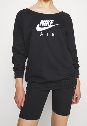 Osoba w czarnej bluzie Nike Air i czarnych dopasowanych szortach stoi na jednolitym białym tle.