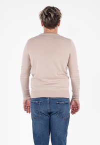 Hombre con cabello corto y iluminado, vistiendo un suéter de manga larga beige y jeans azules, de pie de espaldas contra un fondo blanco.