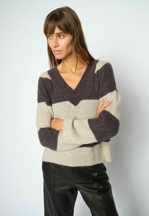 Kvinde med langt brunt hår iført stribet V-hals sweater og sorte bukser, står med korslagte arme og kigger til siden.