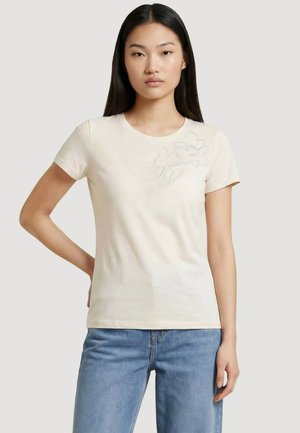 Mujer con cabello largo y negro que lleva una camiseta de manga corta color beige claro con un bordado floral sutil en la parte superior izquierda y jeans azules.