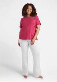 Camisa de manga corta fucsia con bordado floral, combinada con pantalones acampanados blancos y sandalias plateadas, sobre una modelo ante un fondo neutro.