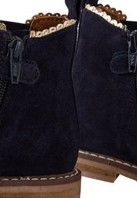 Bottines en suède marine avec fermeture éclair, bord ondulé et talons en bois. Les caractéristiques notables comprennent un matériau texturé et des coutures distinctes.