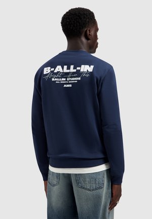 Man draagt een marineblauwe trui met witte tekst "B-ALL-IN" en kleinere tekst op de bovenrug, gecombineerd met vergeelde blauwe spijkerbroek.