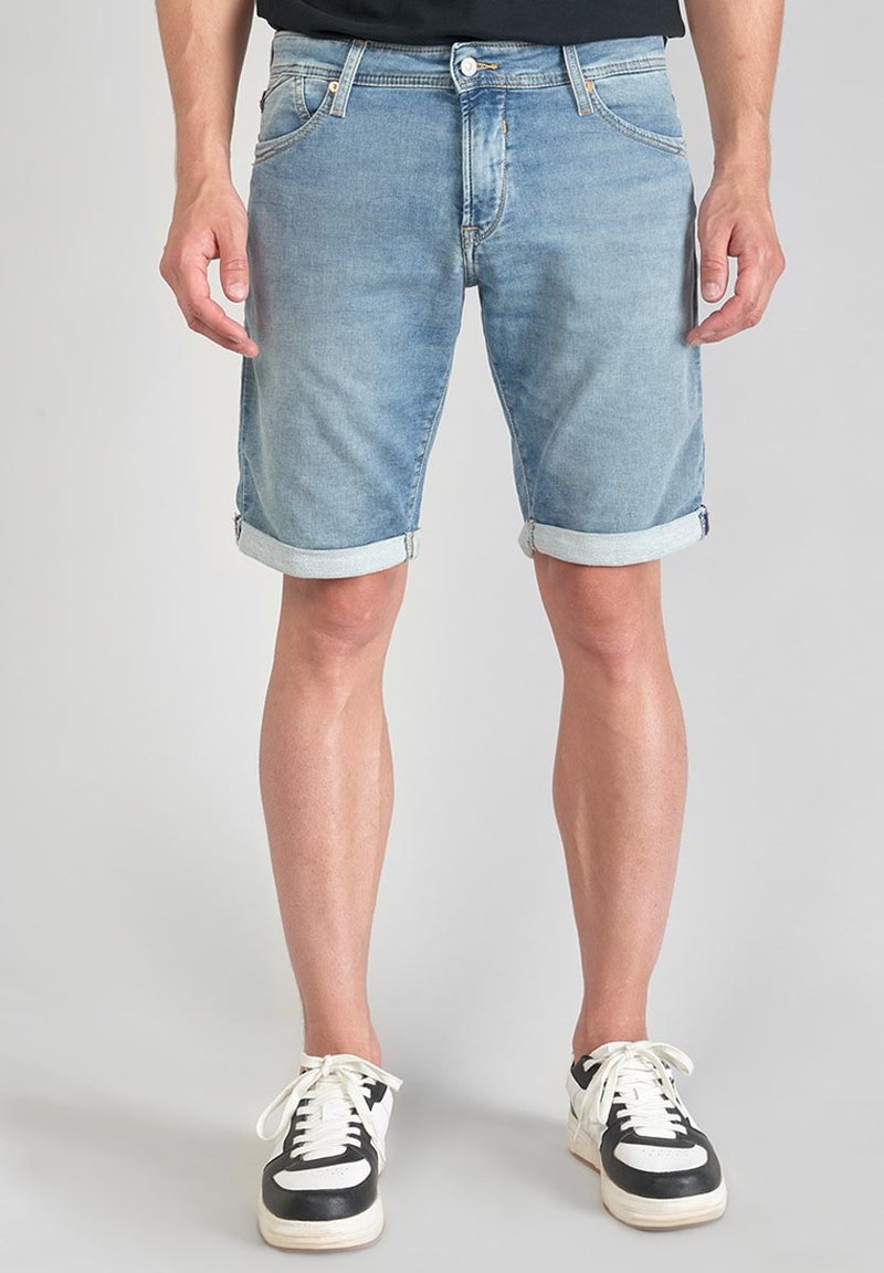Shorts en denim bleu clair avec des ourlets roulés, présentant des poches avant, une fermeture à boutons et des coutures décoratives. Associés à des baskets noir et blanc.