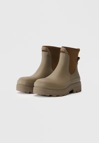 Botas de tobillo impermeables en color beige, con un acabado texturizado, paneles laterales elásticos y una suela gruesa con un patrón para mayor agarre.